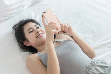 Portre mankeni seksi kadın gri pijama giyiyor, uyuyor ve hafta sonları beyaz yatak odasında akıllı telefon çalıyor.