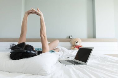 Yeşil pijama giyen kadın bluetooth kulaklıklarla müzik dinliyor. Yatak odasında, hafta sonları beyaz yatakta, bulanık odaklı bulanıklık için.