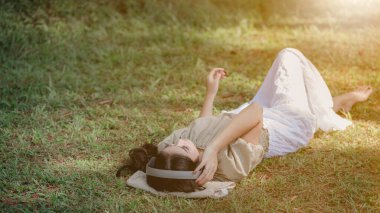 Güzel Asyalı kadın uzanıp kulaklık dinliyor. Çam ormanları bahçesinde bluetooth müzik dinliyor. Açık turuncu işlem geçmişi var.  