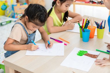 Asyalı çocuklar yaratıcı olmak için renkli kalemler kullanıyor. Okul konseptine dönmek için sınıftaki masaya neşeyle resim çiziyorlar.