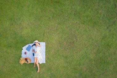 Yukarıdan bakıldığında beyaz elbiseli çekici bir kadın dizüstü bilgisayarla dinleniyor ve yeşil çimlerin arka planında kahve içiyor İHA görüntüsünden.