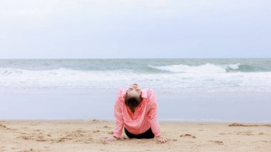 Spor giysi ve pembe ceket giyen genç Asyalı kadın yağmur mevsiminde akşam deniz sahilinde egzersiz yapıyor ve dinleniyor.