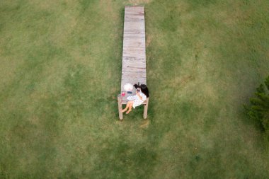 Gökyüzü manzaralı güzel Asyalı kadınlar ve kedi bahçedeki yeşil çimenlerin arkasındaki tahta köprüde dinleniyor ve sıcak kahve içiyorlar.,