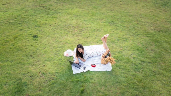 年轻的亚洲女人躺在绿色的玻璃上放松 在花园里用红色的咖啡杯在笔记本电脑上工作 商业技术和自由职业概念 无人机的航空视图 — 图库照片