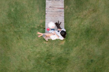 Gökyüzü manzaralı güzel Asyalı kadınlar ve kedi bahçedeki yeşil çimenlerin arkasındaki tahta köprüde dinleniyor ve sıcak kahve içiyorlar.,