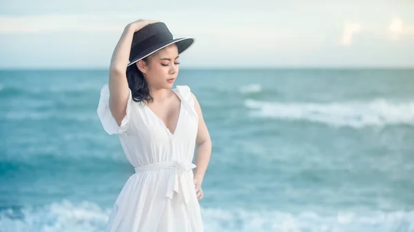 身穿白衣 头戴黑帽的美丽的亚洲女人在海滩上拍照 海浪背景暑假则是周末旅游理念和淡淡的画风 — 图库照片