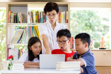 Bir grup Asyalı Öğrenci Okuma ve Bilgisayarı Okuma ve Bakma Masasında, Okul Kütüphanesinde, Kitap Rafları Arkaplanda, Asya Çocuk Eğitimi Yaz Sınıfı Tayland Konsepti ile bayan öğretmenle birlikte. 