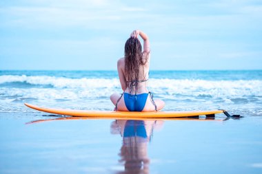 asian girl sitting back posing with her surfboard on the beach in summer blue tone process,