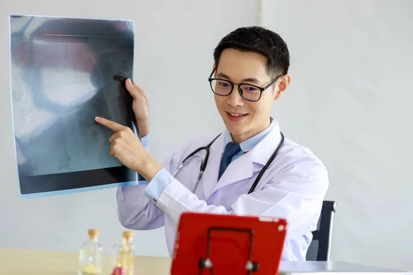asian man doctor showing  x-ray lap sheet and explain the details to patient via video call system in office, thailan