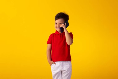 Kırmızı polo tişörtlü çocuk, beyaz şort ve cep telefonlu sarı arka planlı stüdyo fotoğrafı..