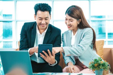 İş çifti dizüstü bilgisayarla birlikte oturuyorlar ve pencere arkaplanının yakınındaki tablet üzerindeki çalışmaları kontrol ediyorlar.