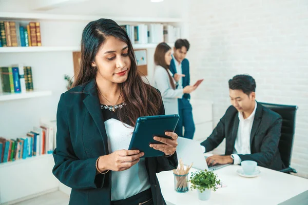 Portrait of a beauty indian business woman CEO smiling. Portrait of a positive looking young business professional standing holding tablet with co-workers talking in the background.