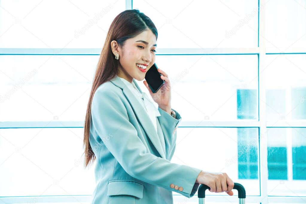 Young businesswoman in a suit pulling a suitcase and talking on the cell phone in front of an airport window