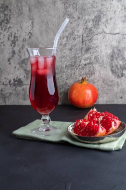 Uzun bir bardak buzlu kırmızı meyve suyu ve gri arka planda pipetler, olgun bir nar meyvesinin yanında. Dikey konum, boşluğu kopyala