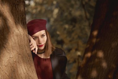 Pretty girl leaning against a tree in the woods, looking away. Wearing black coat and red sweater.
