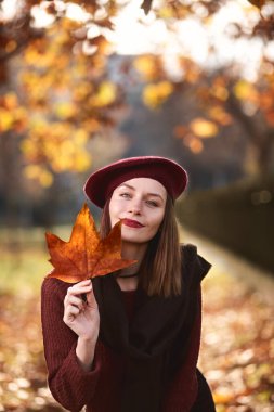Brunette girl smiling and holding a maple leaf in front of her face. Wearing a beret and scarf