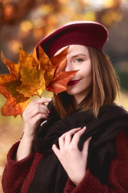 Closeup portrait of young girl, wearing a french beret. Holding maple leaves in her hand, covering her eye