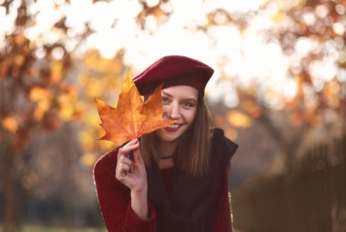 Smiling woman holding in her hands yellow maple leaves covering her eye. Wearing a beret