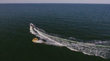Karadeniz 'de motorlu bir tekne tarafından çekilen şişme yüzüğün havadan çekilen görüntüsü. Neşe dolu kuş bakışı beyaz motorlu teknenin, Karadeniz' de insanlarla birlikte şişme halkayı çekerek güneşli bir yaz gününde dalgalanan dalgaların izlerini bırakması..