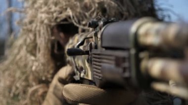 Kamuflajlı bir asker elinde makineli tüfekle karada yatıyor ve baharda nişan alıyor. Kamuflajlı profesyonel bir askere yakından saldırıyor. Çimlere uzanmış bir miğferi var..