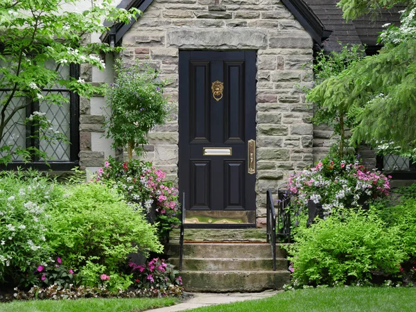 Kurşunlu cam pencereli, çiçeklerle ve çalılarla çevrili taş yüzlü evin ön kapısı.