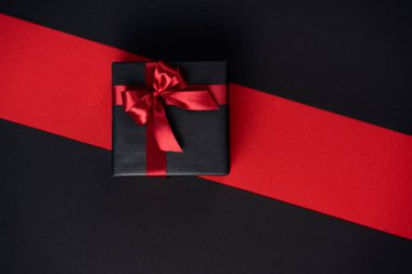 Kırmızı kurdeleli siyah kağıda sarılı bir hediye kutusu ve siyah-kırmızı bir zemin üzerinde yay.