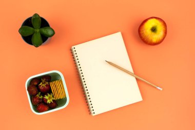 Ofisteki işyerinde sağlıklı atıştırmalıklar. Kalemli defter, ev çiçeği ve turuncu arka planda çilek ve kurabiyeli kap.