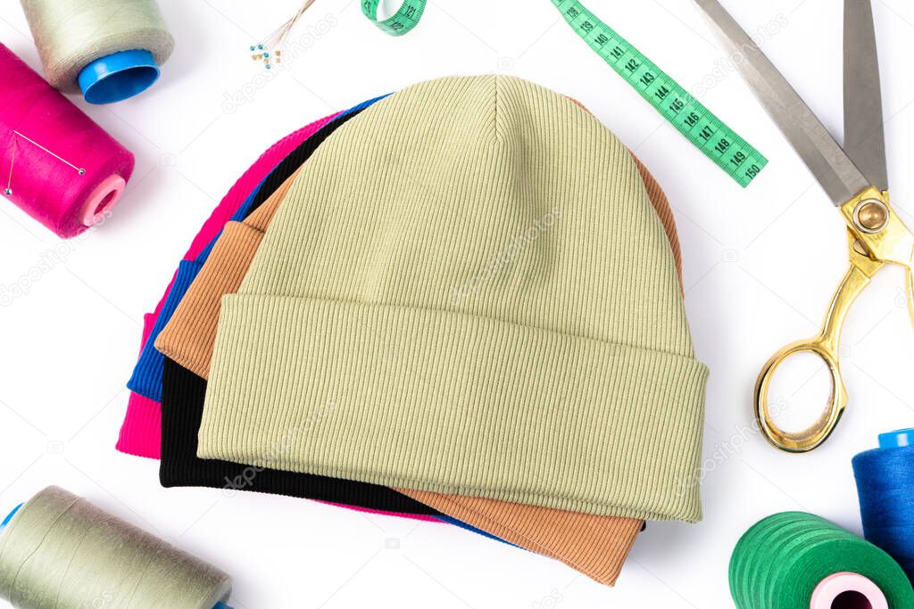 Top view of a stack of insulated stylish beanie hats in different colors with scissors, threads and needles. Tailoring concept