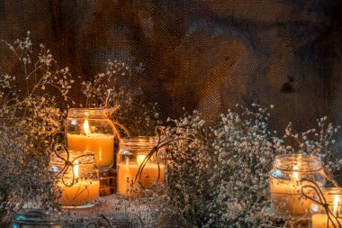 Kavanozda el yapımı yanan beyaz mum kaba tekstil ışıltısı alevli kuru buket çıngıraklı çiçek kırsal karanlık ruh hali turuncu açık mavi