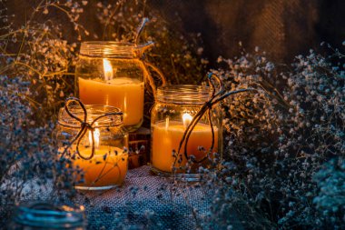 Kavanozda el yapımı yanan beyaz mum kaba tekstil ışıltısı alevli kuru buket çıngıraklı çiçek kırsal karanlık ruh hali turuncu açık mavi