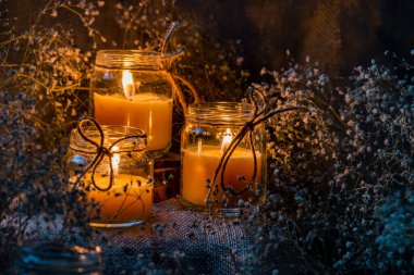 Kavanozda el yapımı yanan beyaz mum kaba tekstil ışıltısı alevli kuru buket çıngıraklı çiçek kırsal karanlık ruh hali turuncu açık mavi