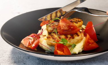 Bıçak ve çatal tabakta hindi pirzolası, patates püresi ve domates var.