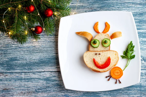 A perky bull from white bread, carrots, cucumbers, parsley as a Breakfast decoration for a child for Christmas and New year on a large white plate on a blue wooden background. — Stock Photo, Image