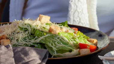 Sezar salatası bir tabak marul, vişneli domates, kraker, parmesan peyniri, ızgara tavuk ve masada siyah bir tabak ve peçete üzerine kaba.