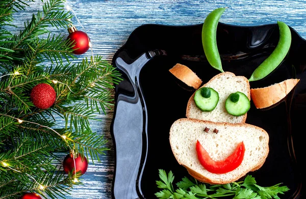 Creative Breakfast sandwich of peas, tomatoes, parsley, cloves, spices, cucumber in the form of a bulls head with large horns on a blue table and fir branches with red balls. — Stock Photo, Image