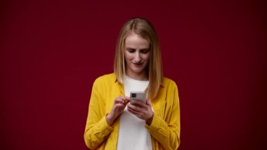 Kız telefondaydı ve aniden bir milyon kazandı, hoş bir şekilde şaşırdı, yumruğunu sıktı, beyaz tişört ve sarı gömlek giydi, arka plan kırmızısı.
