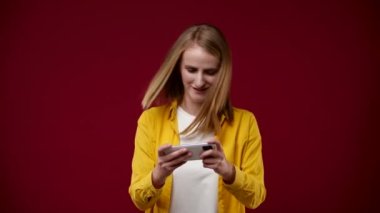 Bir kız telefonda video oyunu oynuyor ve kazanıyor, gülümsüyor, yumruklarını sallıyor, çok mutlu, sarışın, beyaz tişört giyiyor ve sarı gömlek giyiyor, arka plan kırmızı.