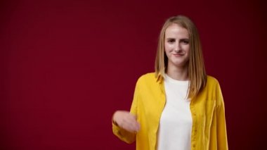 Kız avucunu açık tutuyor ve parmağıyla işaret ediyor, gülümsüyor, beyaz tişört ve sarı gömlek giyiyor, arka plan kırmızı, kız sarışın.