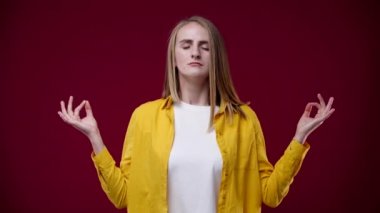 Bir kız kameranın önünde duruyor ve meditasyon yapıyor, kollarını yana doğru açıyor ve düşünüyor, beyaz tişört ve sarı gömlek giyiyor, arka plan kırmızı.