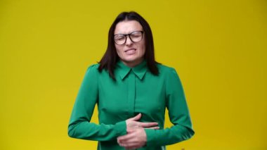 Kadının midesi çok kötü ağrıyor, üzgün, ellerini karnına koyuyor, gözlük takıyor, esmer, yeşil gömlek giymiş, arka plan sarı.