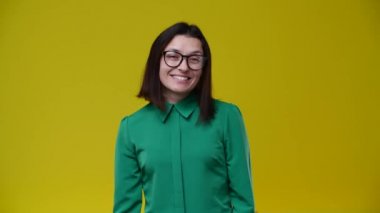Esmer gülüyor ve ellerini göğsüne ve yanaklarına koyuyor, neşeli, gözlük takıyor, kısa saçlı, yeşil gömlekli, arka plan sarı.