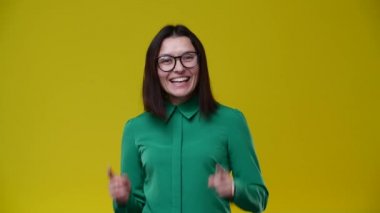 Kız kameraya senin en iyisi olduğunu gösteriyor. Gözlük takıyor, esmer, yeşil gömlek giyiyor. Arka plan sarı.