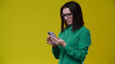 Kız kaseti çeviriyor ve çok şaşırıyor, gözlük takıyor, esmer, yeşil gömlek giymiş, arka plan sarı.