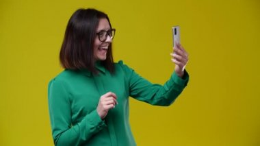 Kız bir video görüşmesinde konuşuyor, elini sallıyor ve gülümsüyor, esmer, gözlük takıyor, yeşil gömlek giyiyor, arka plan sarı.