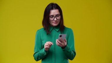 Esmer kadın bir telefon aldı ve çok iyi bir haber olduğunu söyledi. Yeşil bir gömlek giymiş, arka planı sarı, gözlük takıyor ve kısa saçlı.