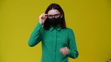 Gözlüklü bir kadın kameraya bakar ve maskeyi çıkarır, gülümser, maske siyahtır, yeşil gömlek giyer, arka plan sarıdır.