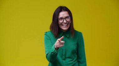 Kız kameraya bakıyor ve sana gülüyor, esmer, kısa saçlı, gözlük takıyor, yeşil gömlek giyiyor, arka plan sarı.