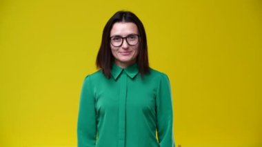 Kız gözlüklerini çıkarır ve göz kırpar, kameraya bakar, yüzüne gülümser, yeşil gömlek ve sarı bir arka plan giyer.