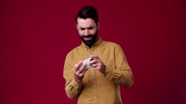 Adam telefonda yarışıyor ve kazanıyor, gülümsüyor, neşeli, ellerini sallıyor, sakallı, siyah saçlı, kahverengi gömlekli, arka plan kırmızısı
