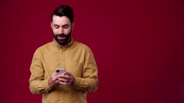 Adam telefonu elinde tutuyor ve yan tarafı işaret ediyor, marka, logo, ürün, boş alan, gülümsüyor, kahverengi gömlek giyiyor, arka plan kırmızı, sakallı, siyah saçlı.